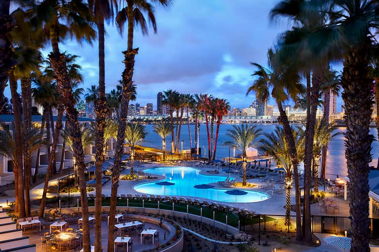 Illuminated resort pool with palm trees overlooking waterfront cityscape at dusk