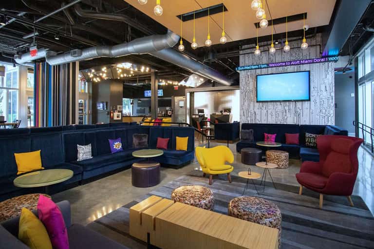Modern hotel lounge with navy blue sectional sofa, colorful pillows, pendant lighting, and casual seating areas.