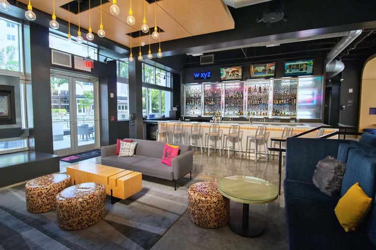 Modern hotel bar and lounge with pendant lighting, gray seating, patterned ottomans, and floor-to-ceiling windows