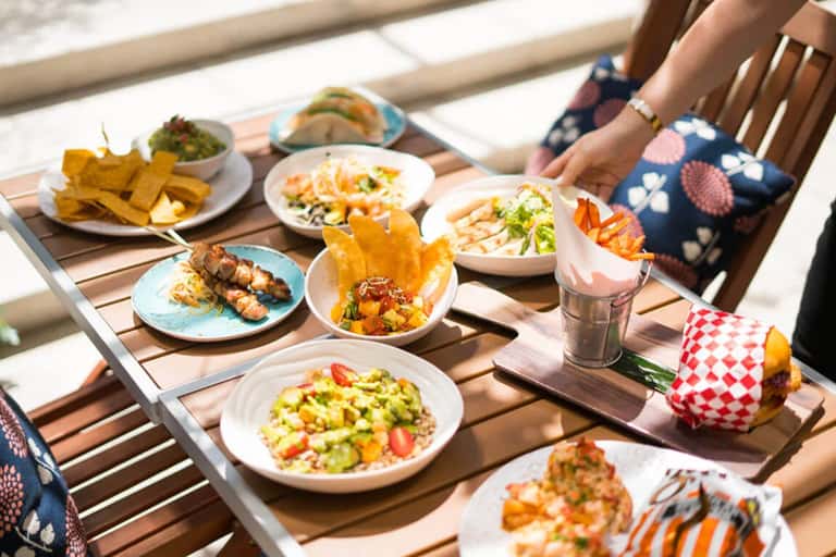 Outdoor dining table with various dishes, appetizers, and beverages in bright sunlight