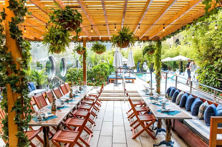 Outdoor dining area with wooden pergola, hanging plants, and red chairs overlooking a resort pool