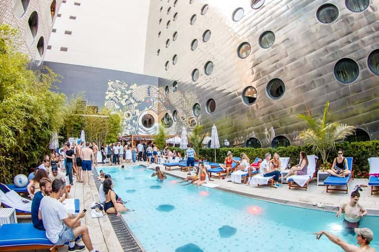 Indoor resort pool surrounded by tall buildings with circular windows and lush tropical plants, filled with guests enjoying the water