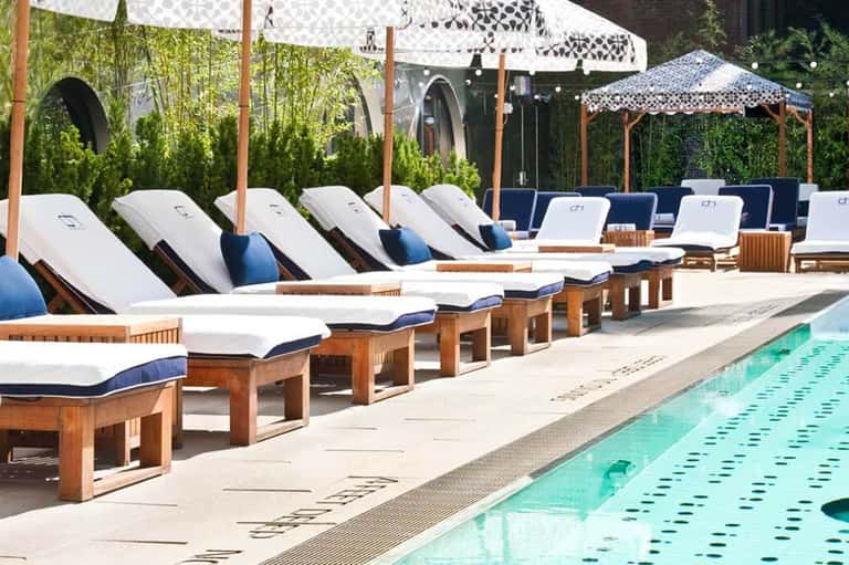 Luxury poolside lounge area with wooden chaise lounges, white cushions, blue pillows, patterned umbrellas, and manicured hedges