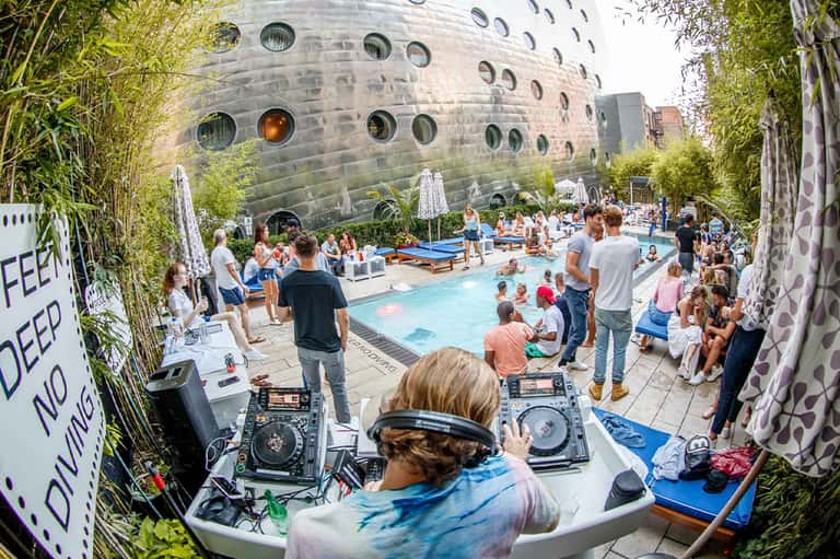 DJ performing at a lively poolside party with guests and a distinctive cylindrical building with circular windows