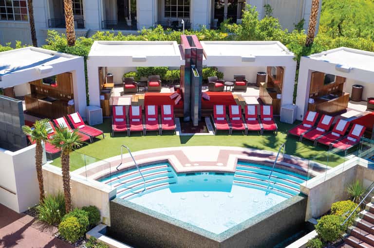 Outdoor hotel pool area with red loungers, white cabanas, and palm trees under sunny sky
