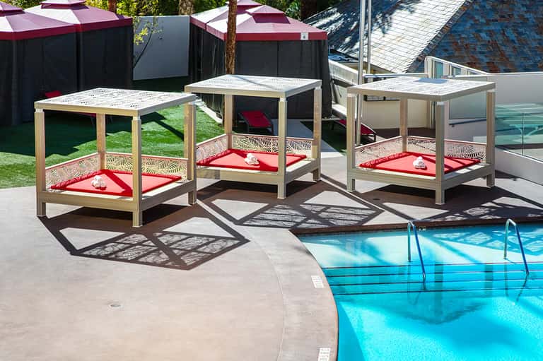 Three wooden cabana beds with red cushions by a swimming pool on a sunny patio