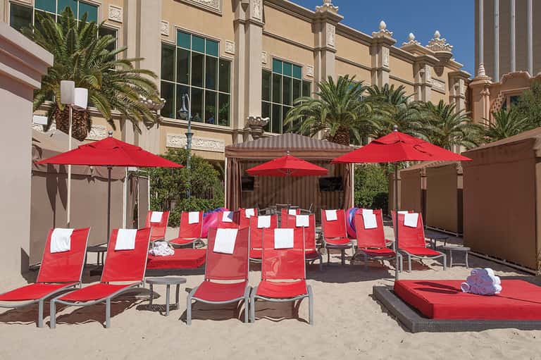 Beach courtyard with red loungers, umbrellas, and palm trees surrounding a sandy area with modern hotel architecture
