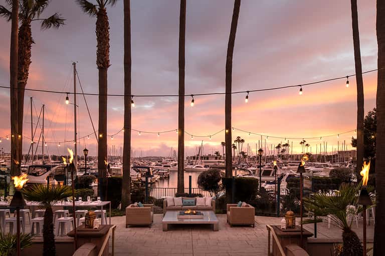Waterfront terrace at sunset with palm trees, string lights, sailboats, and lounge seating overlooking marina