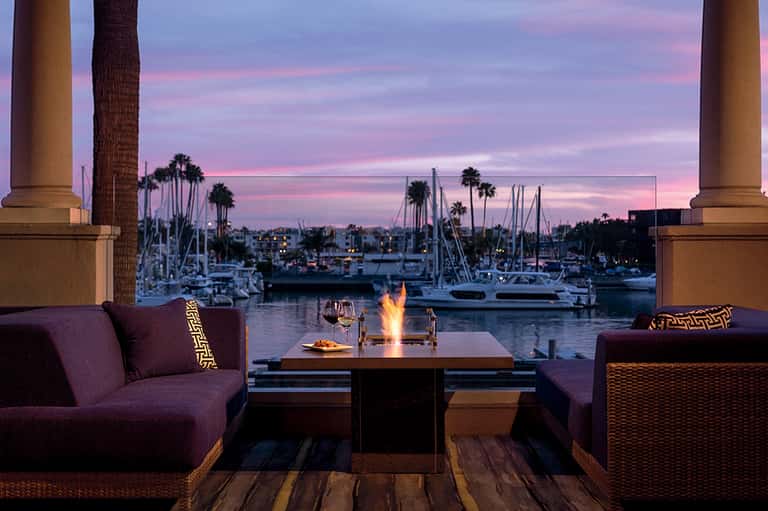 Waterfront lounge at sunset with fire pit, purple seating, and sailboats docked in marina background