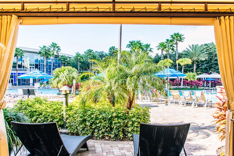 Resort pool area with palm trees, lounge chairs, and yellow cabanas viewed from covered patio