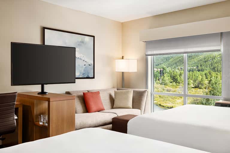 Modern hotel room with two beds, mountain forest view through glass doors, TV and seating area