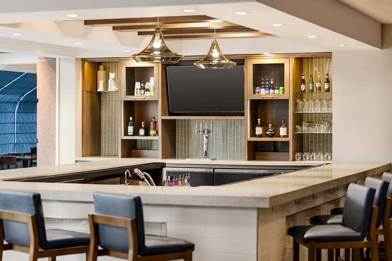 Modern hotel bar with wooden shelving, brass pendant lights, and a curved white counter with bar seating