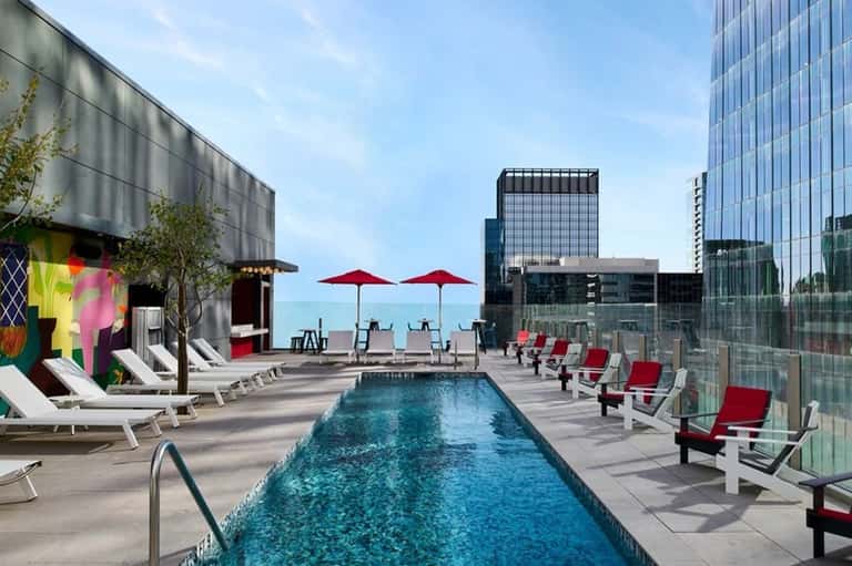 Rooftop pool deck with lounge chairs, red umbrellas, and modern high-rise buildings surrounding the urban pool area