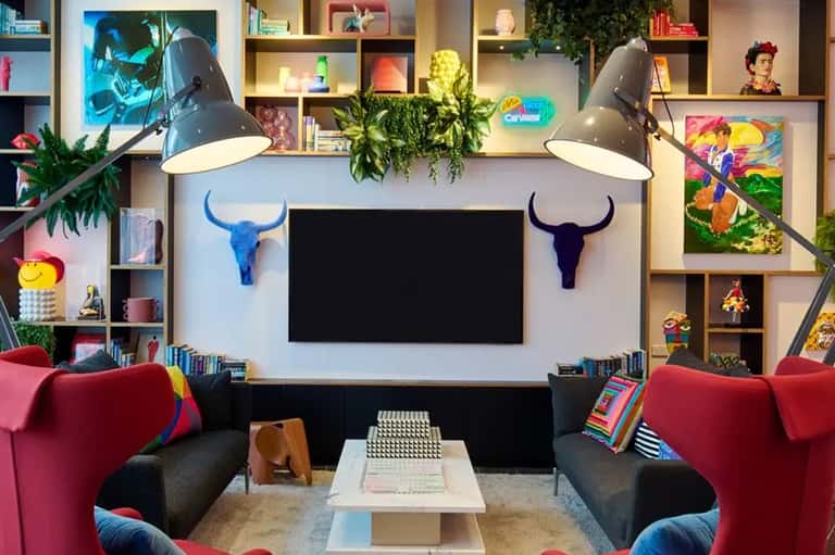 Modern living room with colorful artwork, red armchairs, blue pendant lamps, and shelving unit with TV