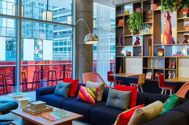 Vibrant modern lounge with colorful pillows on navy sofa, floor-to-ceiling windows overlooking city skyline, and shelving with decorative items