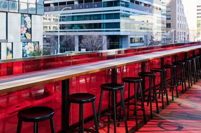Rooftop bar with bright red counter, black bar stools, and urban skyline backdrop with modern buildings