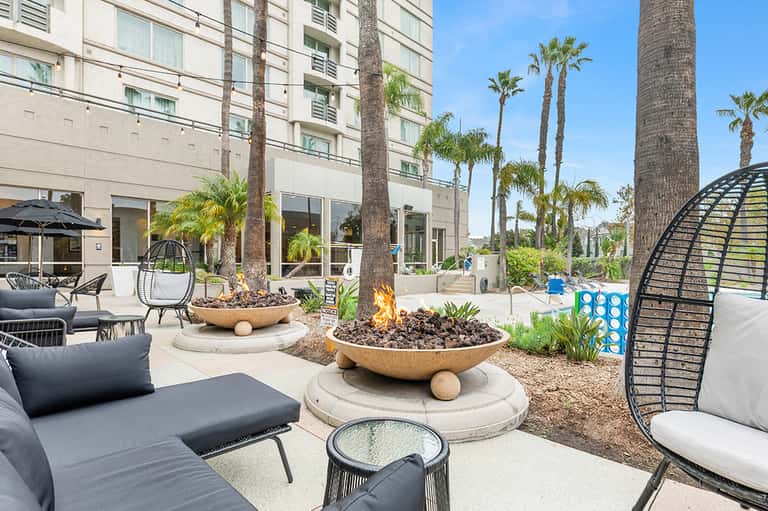 Modern resort courtyard with fire pits, lounge seating, palm trees, and contemporary apartment building
