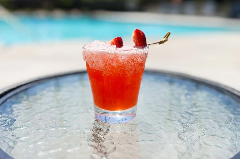 Red cocktail with sugar rim and strawberry garnish on poolside table overlooking blue water
