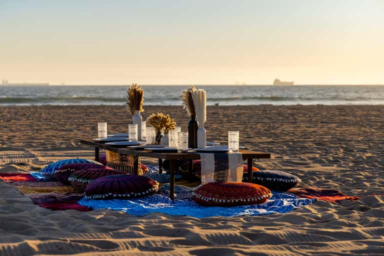Beachfront dinner setup with colorful patterned cushions, candles, and floral centerpieces on sand at sunset