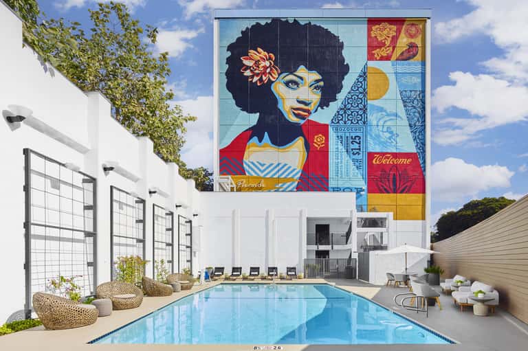 Modern resort pool with colorful portrait mural, white loungers, and grid trellis fence under blue sky