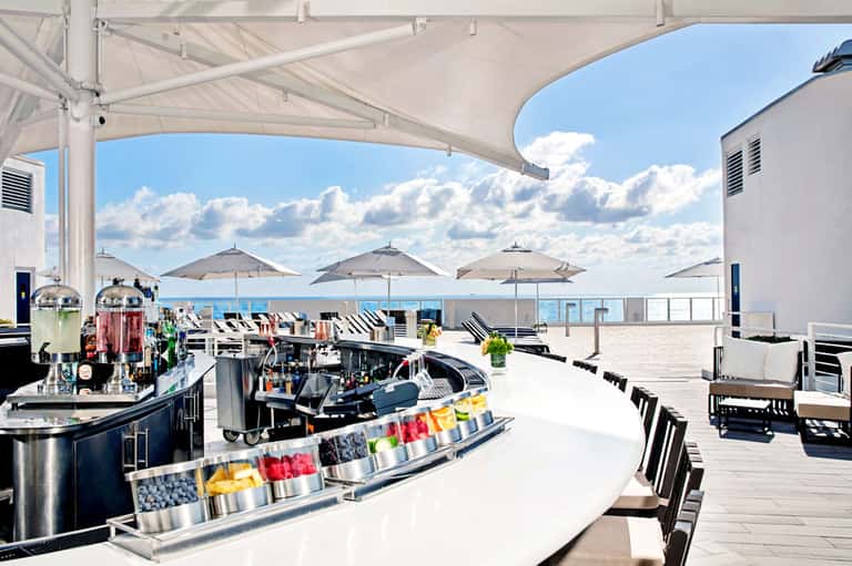 Rooftop bar with white umbrellas, bar counter with colorful garnishes, and ocean views under blue sky