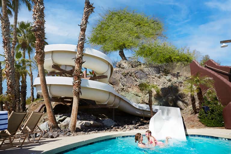 Spiral white water slide at resort pool surrounded by palm trees and lounge chairs