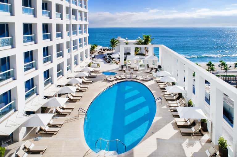 Aerial view of modern beachfront resort with oval pool, loungers, and ocean beyond