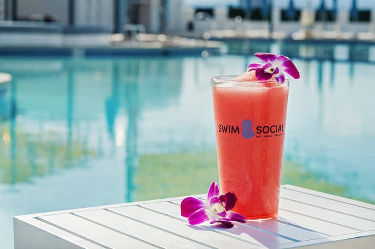 Coral cocktail with orchid flower garnish at poolside bar with turquoise water background