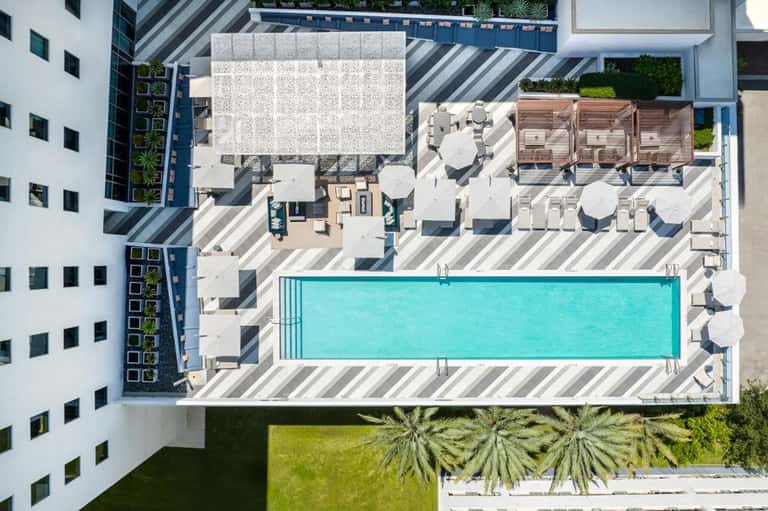 Aerial view of modern rooftop with rectangular pool, lounge chairs, umbrellas, and striped deck surrounded by white buildings