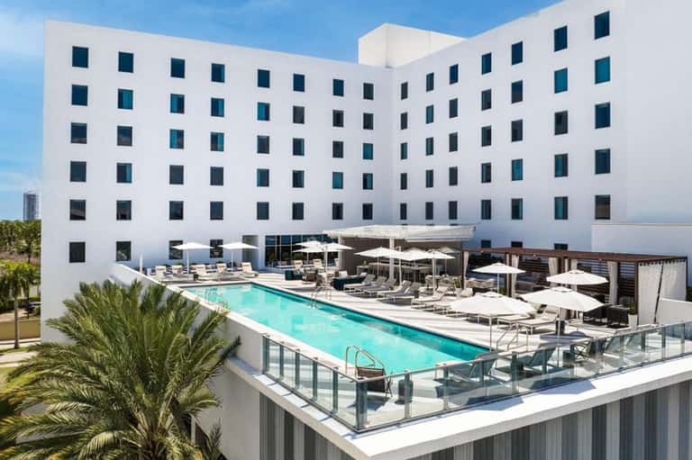 Modern white hotel building with rooftop pool, lounge chairs, and umbrellas overlooking the waterfront