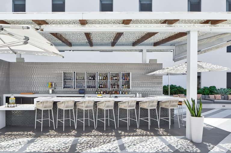 Modern outdoor bar under pergola with white stools, patterned backsplash, and lounge seating in background