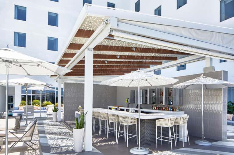 Modern hotel bar and lounge area with white umbrellas, pergola shade structure, and contemporary furnishings