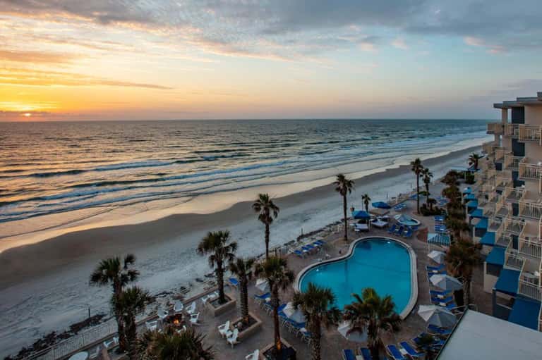 Beachfront resort at sunset with multi-story building, pool, palm trees, and sandy beach with ocean waves