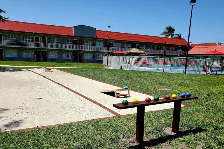 Outdoor cornhole game and bocce ball setup on sandy court with resort building and pool in background