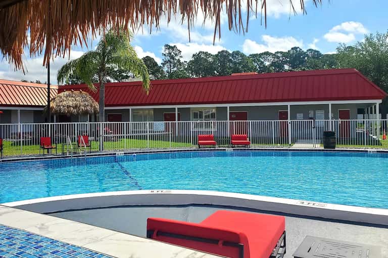 Resort pool with red metal roof building, white railings, lounge chairs, and tropical landscaping under blue sky