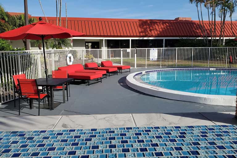 Resort pool area with red lounge chairs, umbrella, and white fencing under clear blue sky