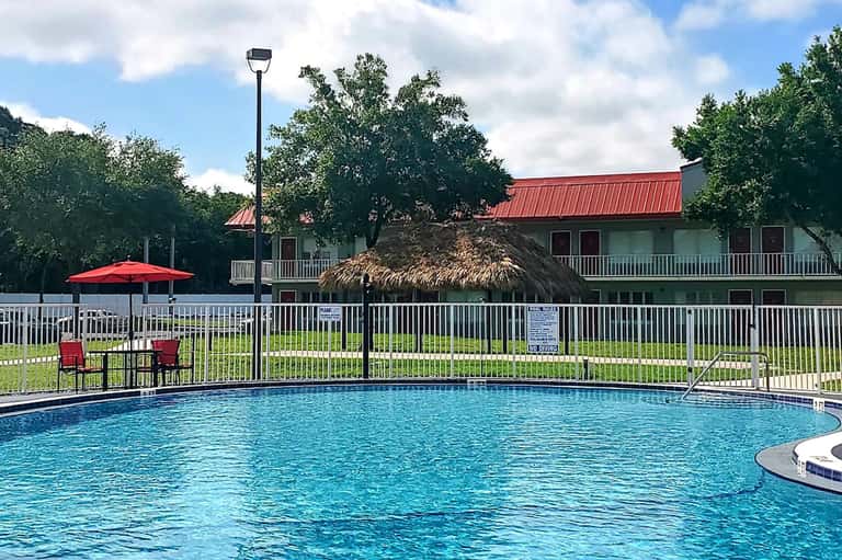 Resort swimming pool with white fence, red umbrella, and thatched-roof cabana surrounded by trees