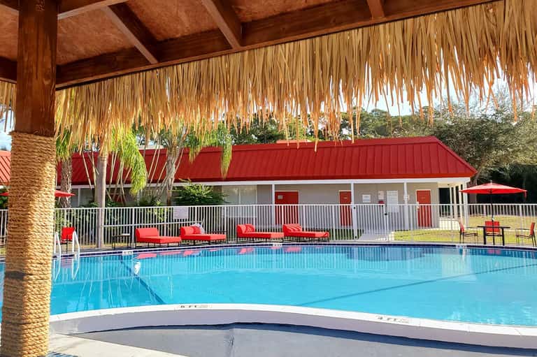 Tropical resort pool with thatched palapa roof, red lounge chairs, and red-roofed building with white railings