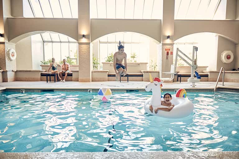 Indoor swimming pool with children playing on inflatable unicorn float, arched windows above