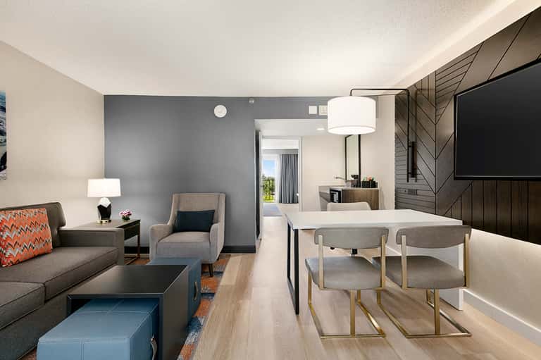 Modern hotel suite with gray accent wall, living area with gray sofa, dining table with chairs, and kitchenette