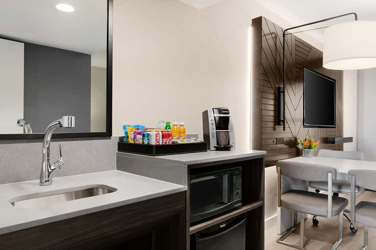 Modern hotel room kitchenette with white countertop, dark cabinetry, sink, microwave, and beverage station with snacks