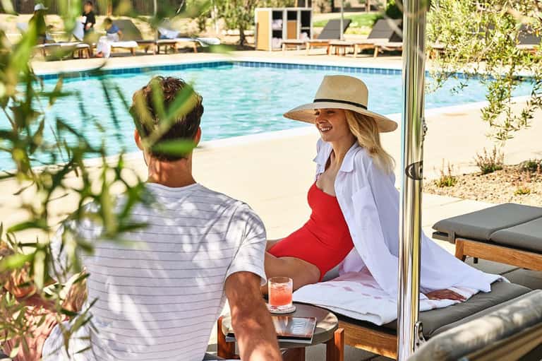 Couple relaxing poolside under pergola with ocean view, woman in red swimsuit wearing straw hat