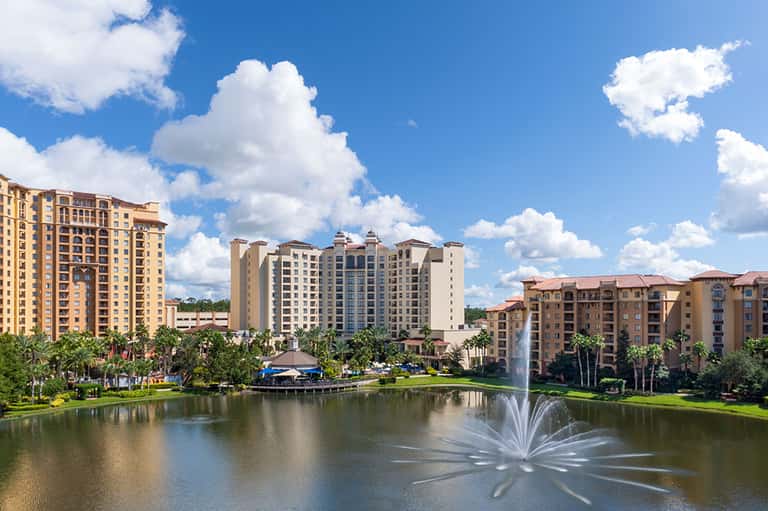 Lakefront resort with multi-story apartment buildings, palm trees, and decorative fountain under blue sky