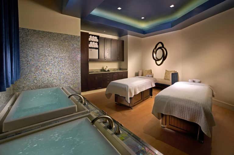 Spa treatment room with two massage beds, hot tubs, and wooden cabinetry with ambient lighting