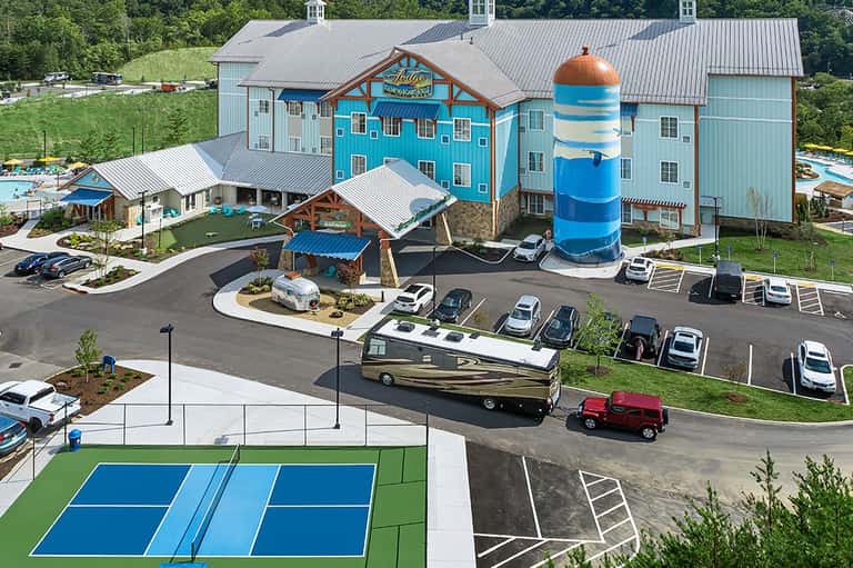 Bright turquoise resort building with striped lighthouse tower, parking area, and tennis courts surrounded by green hills