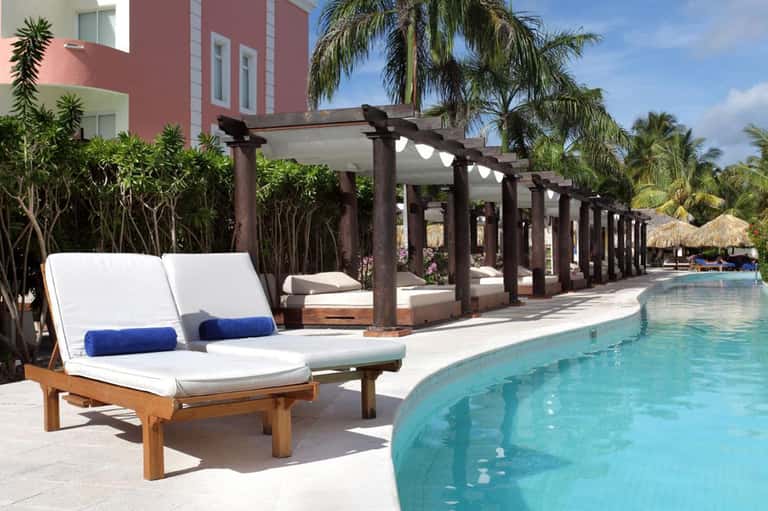 Luxury resort pool area with wooden lounge chairs, pergola structures, and tropical palm trees