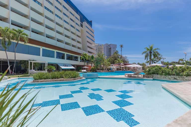 Resort pool with blue and white checkered tile pattern, palm trees, and multi-story hotel building under clear sky