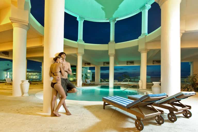 Couple by illuminated indoor pool with columns and night city views through curved windows
