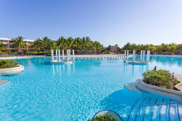 Large resort pool with blue water, white columns, and palm trees under clear sky