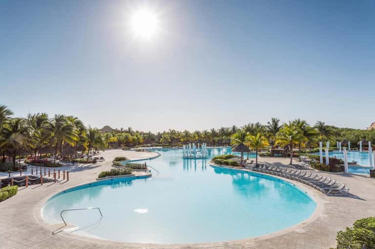 Expansive resort pool complex with curved sections, loungers, palm trees, and blue umbrellas under clear sky